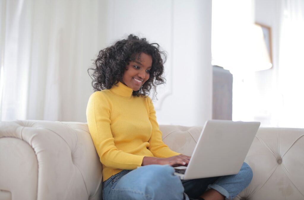Positive young ethnic lady in casual yellow sweater and jeans sitting on comfortable couch and working on laptop while spending time at home